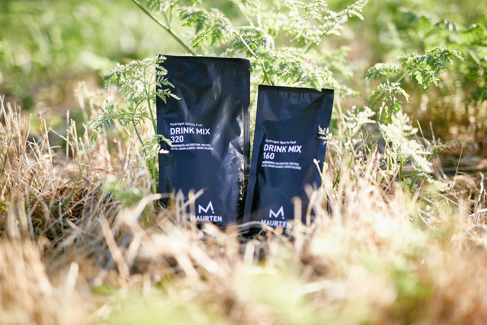 Two black pouches of Maurten drink mix, labeled 320 and 160, stand upright on dry grass. Green foliage surrounds them, giving a natural, fresh feel.